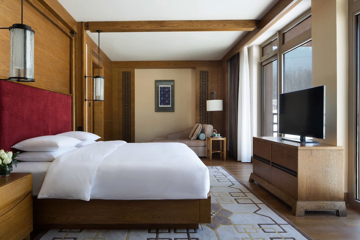 A hotel room with a large bed, wooden headboard, a TV, a desk with a lamp, and a window with a view of trees.