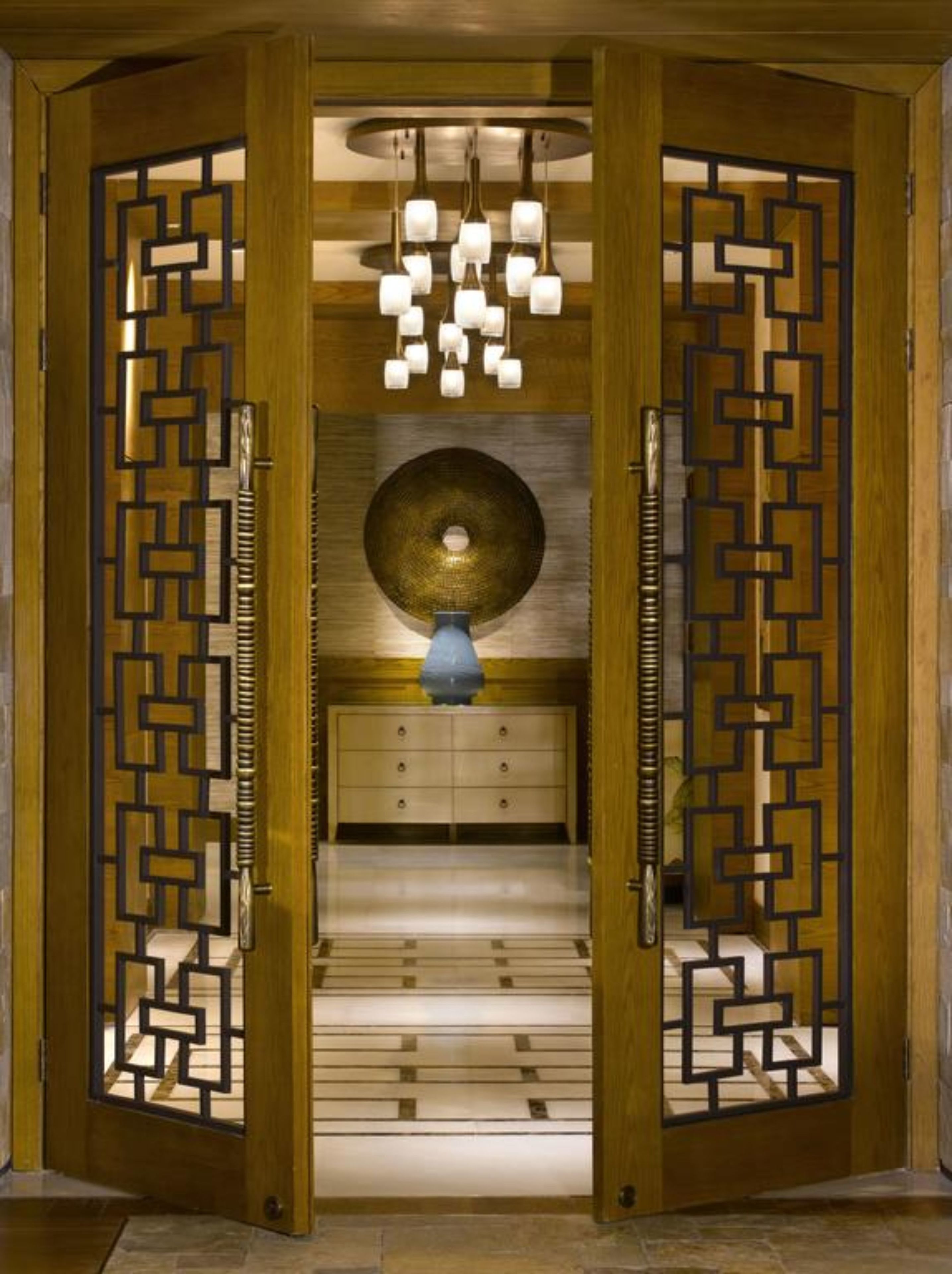 Una habitación con puerta de madera con diseños geométricos, un candelabro central, un objeto decorativo redondo en la pared y un florero azul sobre un mueble.