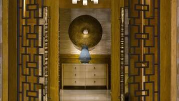 Una habitación con puerta de madera con diseños geométricos, un candelabro central, un objeto decorativo redondo en la pared y un florero azul sobre un mueble.