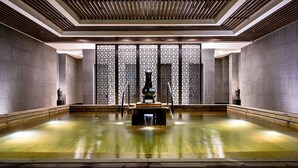 Una amplia piscina interior con una fuente central, rodeada por paredes altas y texturizadas y un techo de listones.