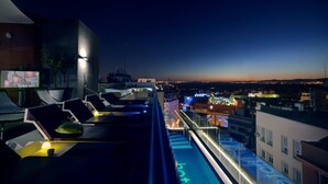 Un salon sur le toit, doté de chaises longues et d’une piscine, offrant une vue sur le paysage urbain au crépuscule.