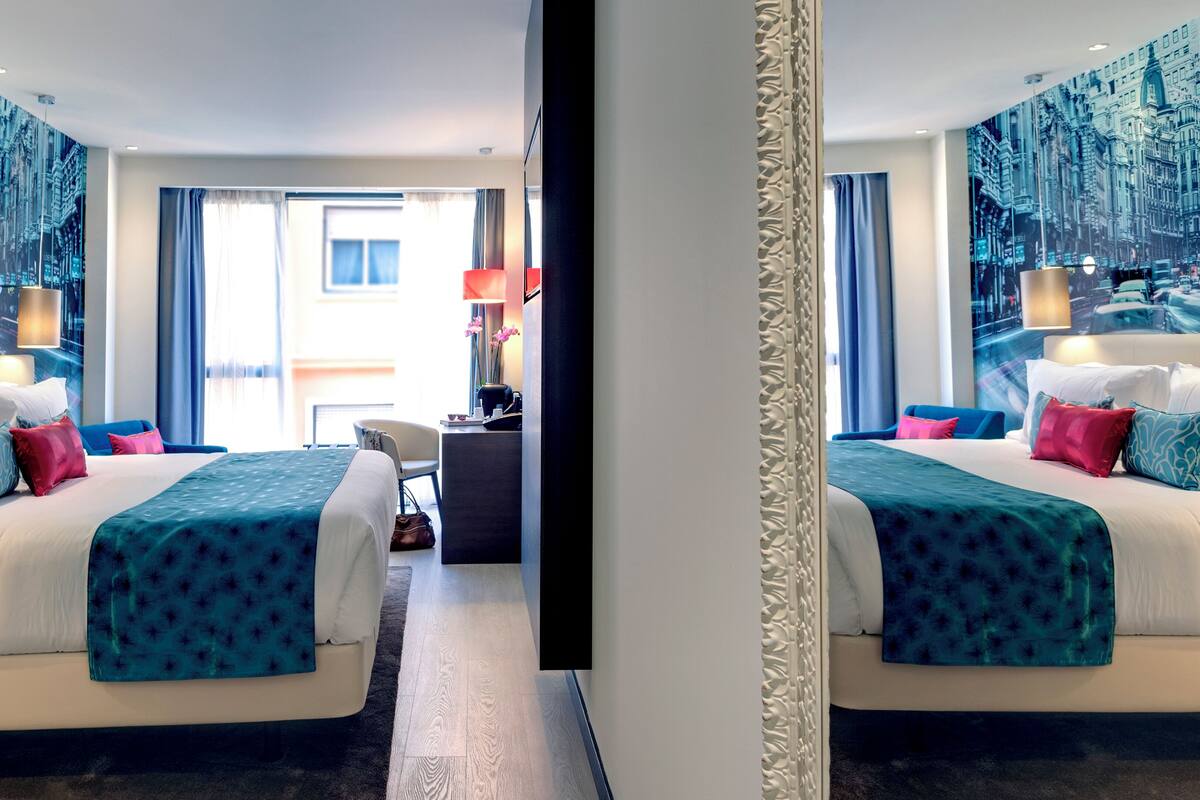 Habitación de hotel con dos camas, un escritorio y vista panorámica de la ciudad a través de amplios ventanales.