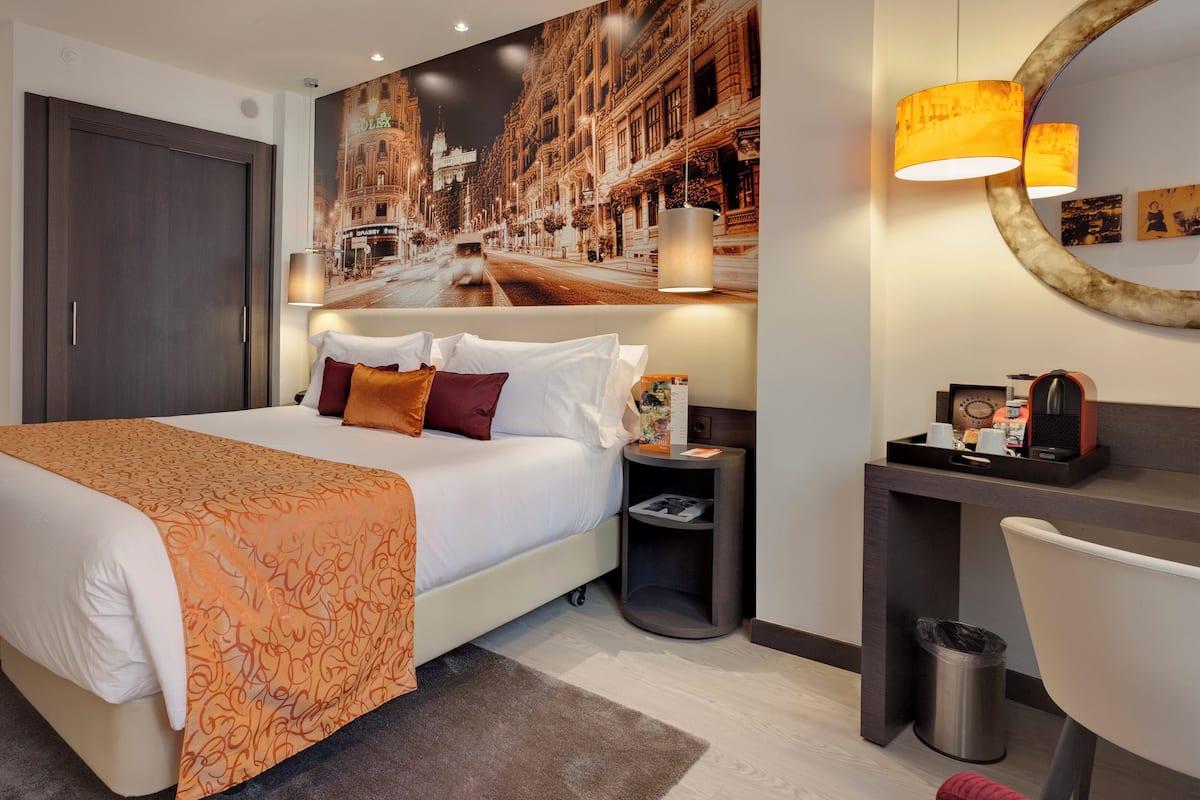 Ein Hotelzimmer mit einem großen Bett, einem Schreibtisch mit Stuhl, einem Mülleimer und einer Wandlampe.