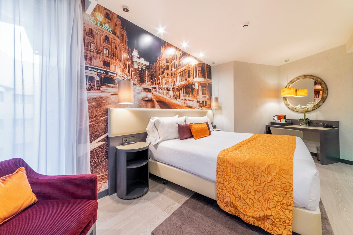 Ein Hotelzimmer mit einem großen Bett, einem Schreibtisch, einem Sessel und Blick auf eine Stadtstraße durch ein Fenster.
