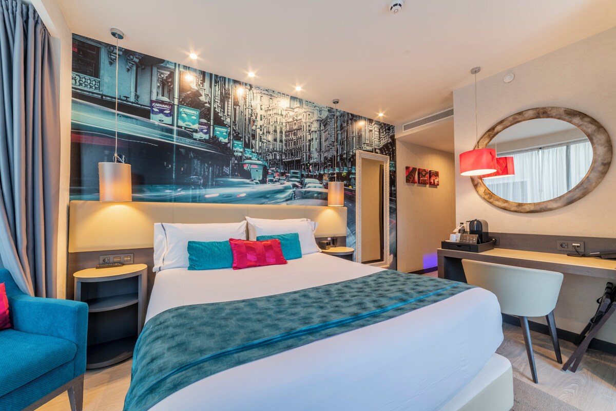 Ein Hotelzimmer mit einem großen Bett, einem Schreibtisch, einem Sessel, einem runden Spiegel und einem Stadtbild-Wandgemälde.