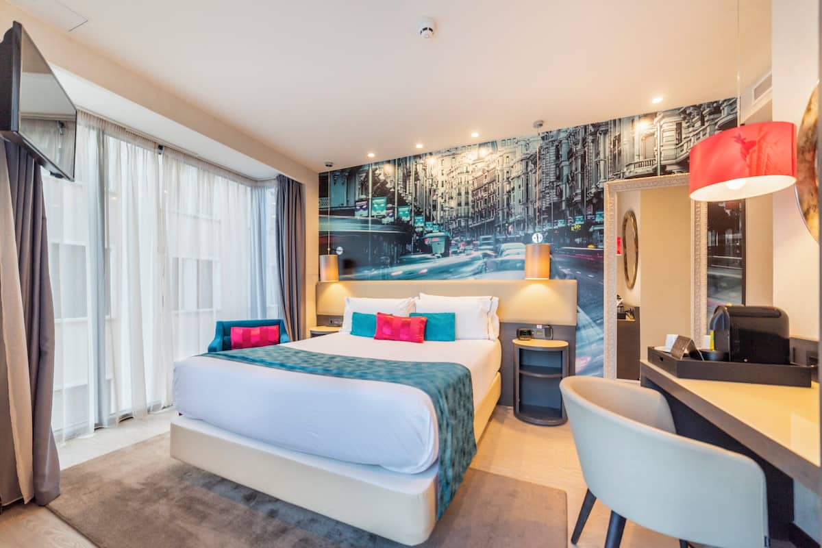Ein modernes Hotelzimmer mit einem großen Bett, einem Schreibtisch, einem Sessel und einem Wandgemälde mit Stadtansicht.
