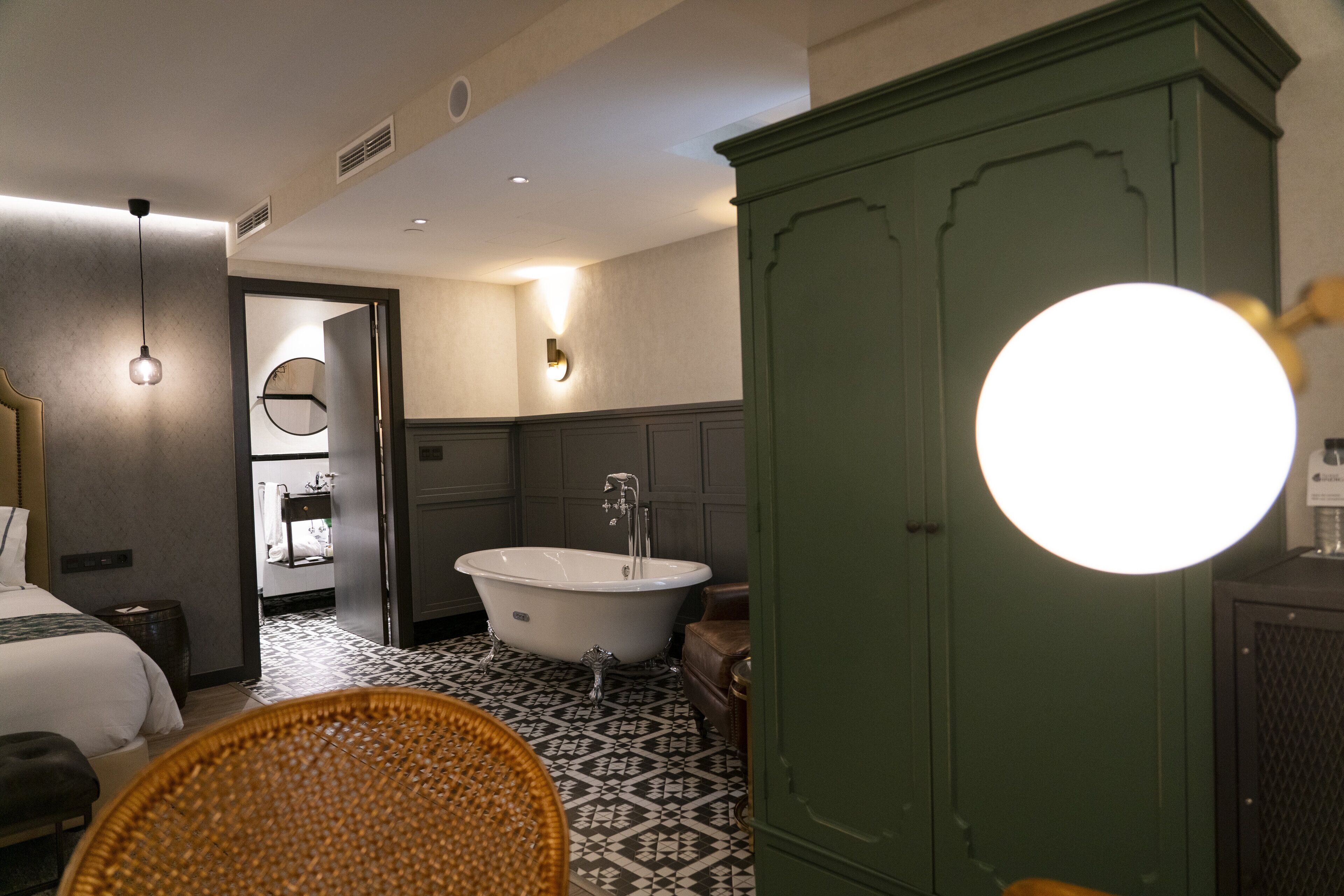 A hotel room with a large green wardrobe, a clawfoot bathtub, a round mirror, and a bed with a headboard.