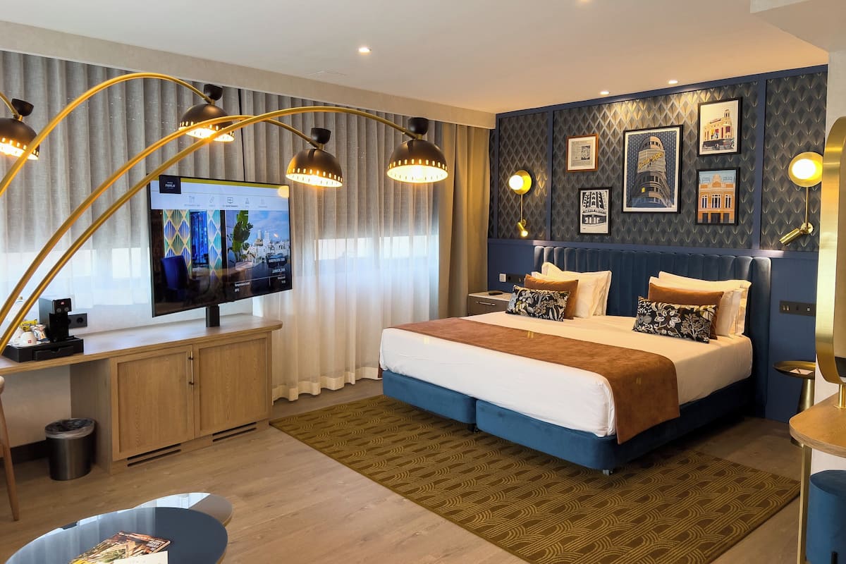Ein modernes Hotelzimmer mit einem großen Bett, einem Schreibtisch mit Computer und einem Fernseher, der eine Stadtansicht zeigt.