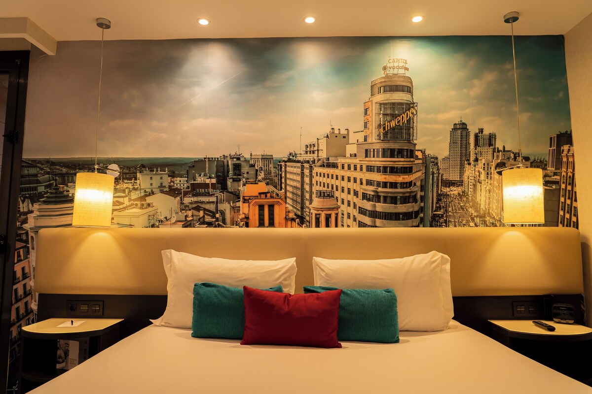 Habitación de hotel con un gran mural de paisaje urbano en la pared, una cama con almohadas blancas y turquesas, y dos lámparas de noche.