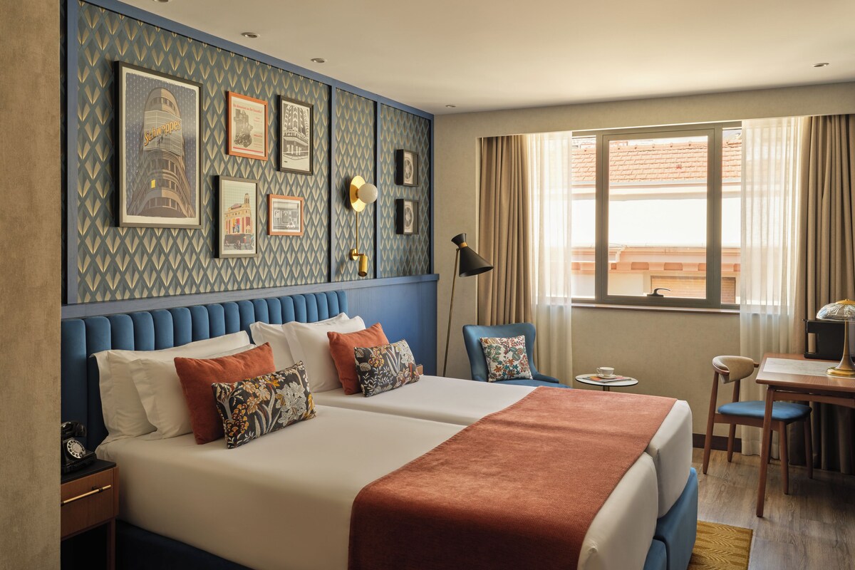 A hotel room with a large bed, a desk, a chair, and a window with curtains.