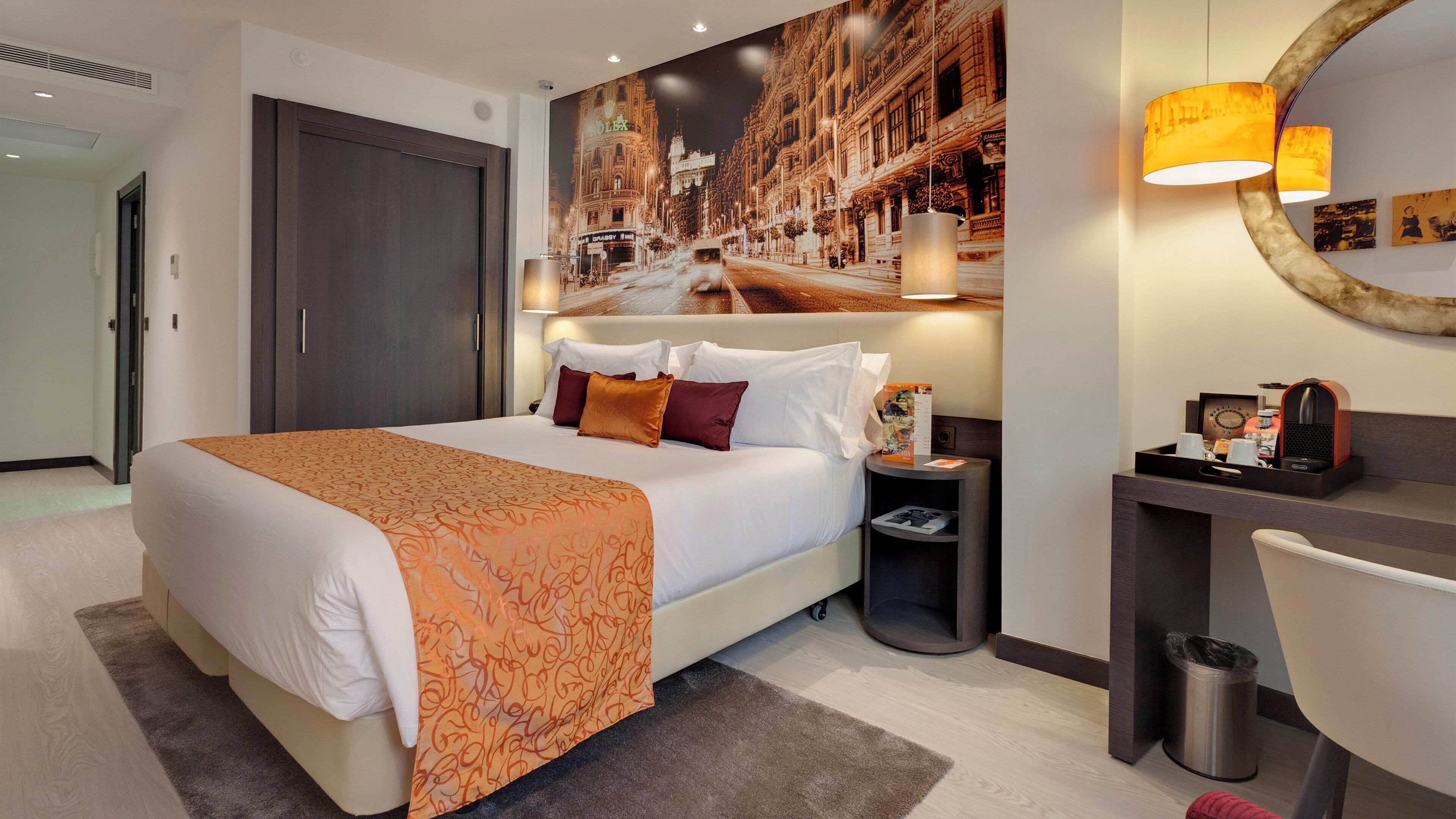 A hotel room with a large bed, a desk with a chair, a trash bin, and a wall-mounted lamp.