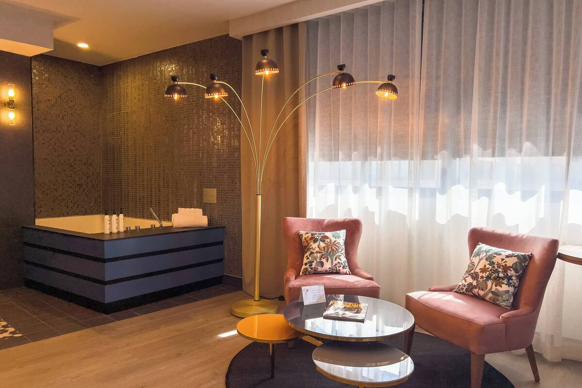 Un baño moderno con una bañera grande, un tocador con artículos de aseo, y un espejo. Hay dos sillones rosas con cojines estampados, una pequeña mesa redonda y una lámpara de pie alta.