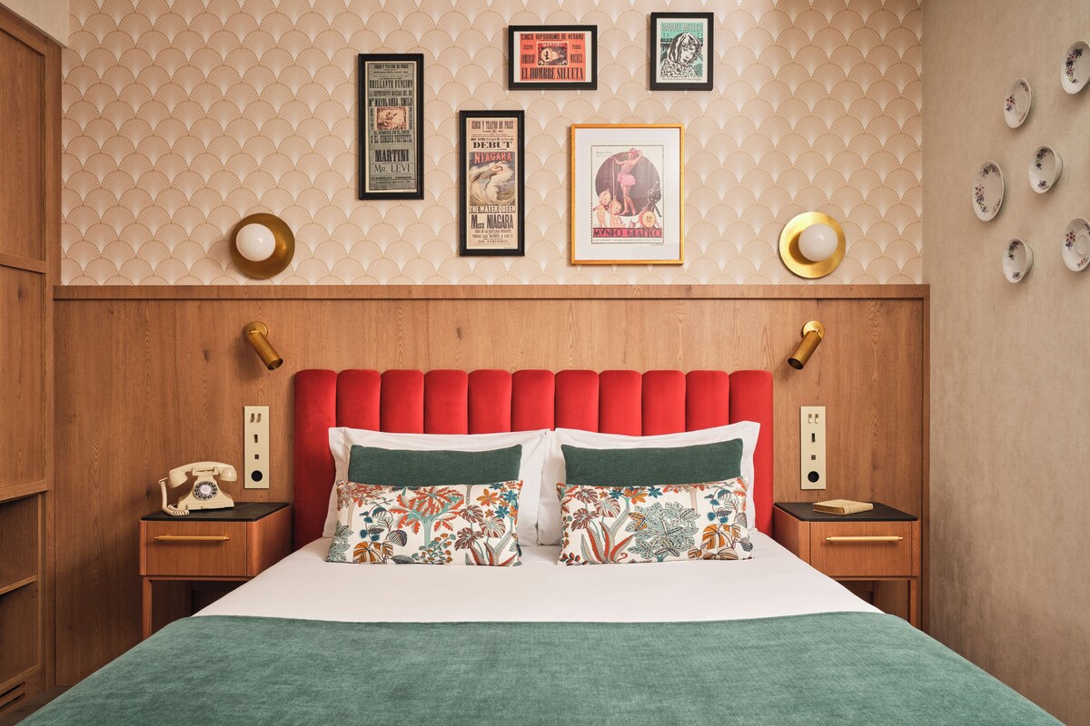 A hotel room with a classic design, featuring a wooden headboard, a bed with floral-patterned pillows, and a wall adorned with vintage posters.