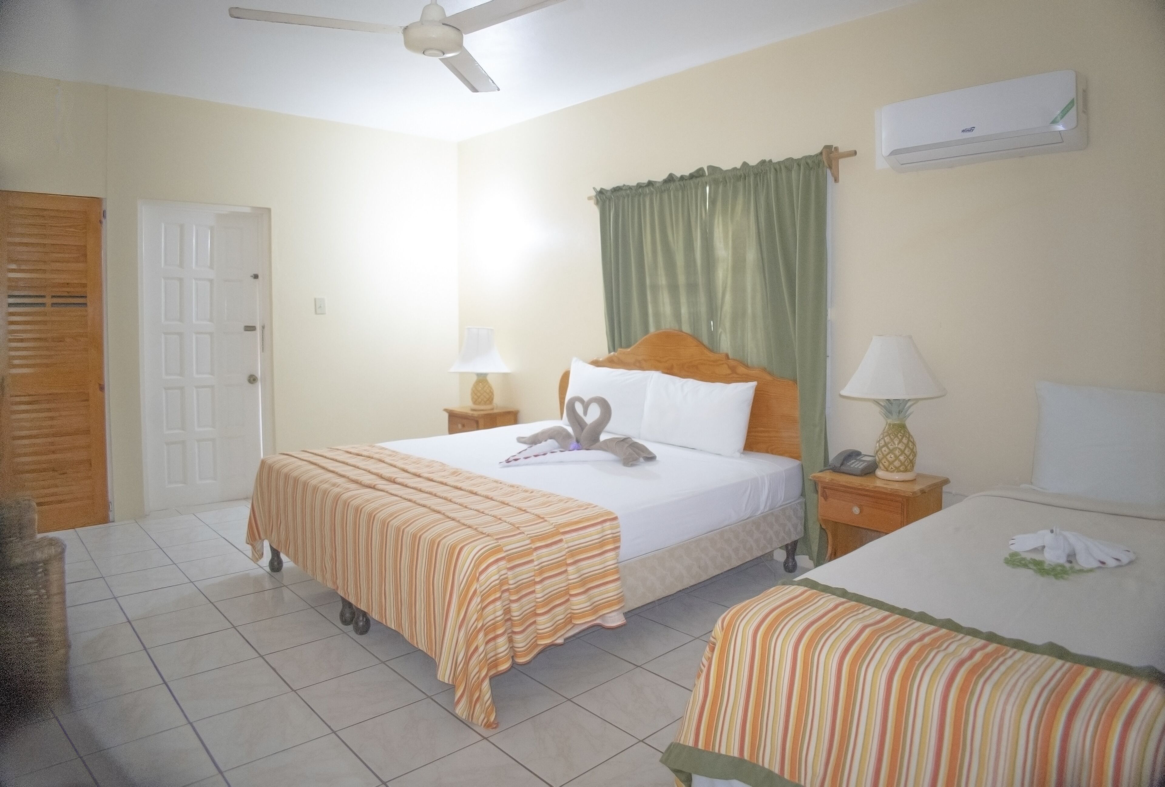 Habitación de hotel con cama, mesita de noche, lámpara, ventilador de techo y aire acondicionado, además de una puerta.