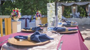 Een gedekte tafel voor een formeel evenement buiten, met een rode tafelkleed, blauwe servetten en glazen vazen met bloemen.