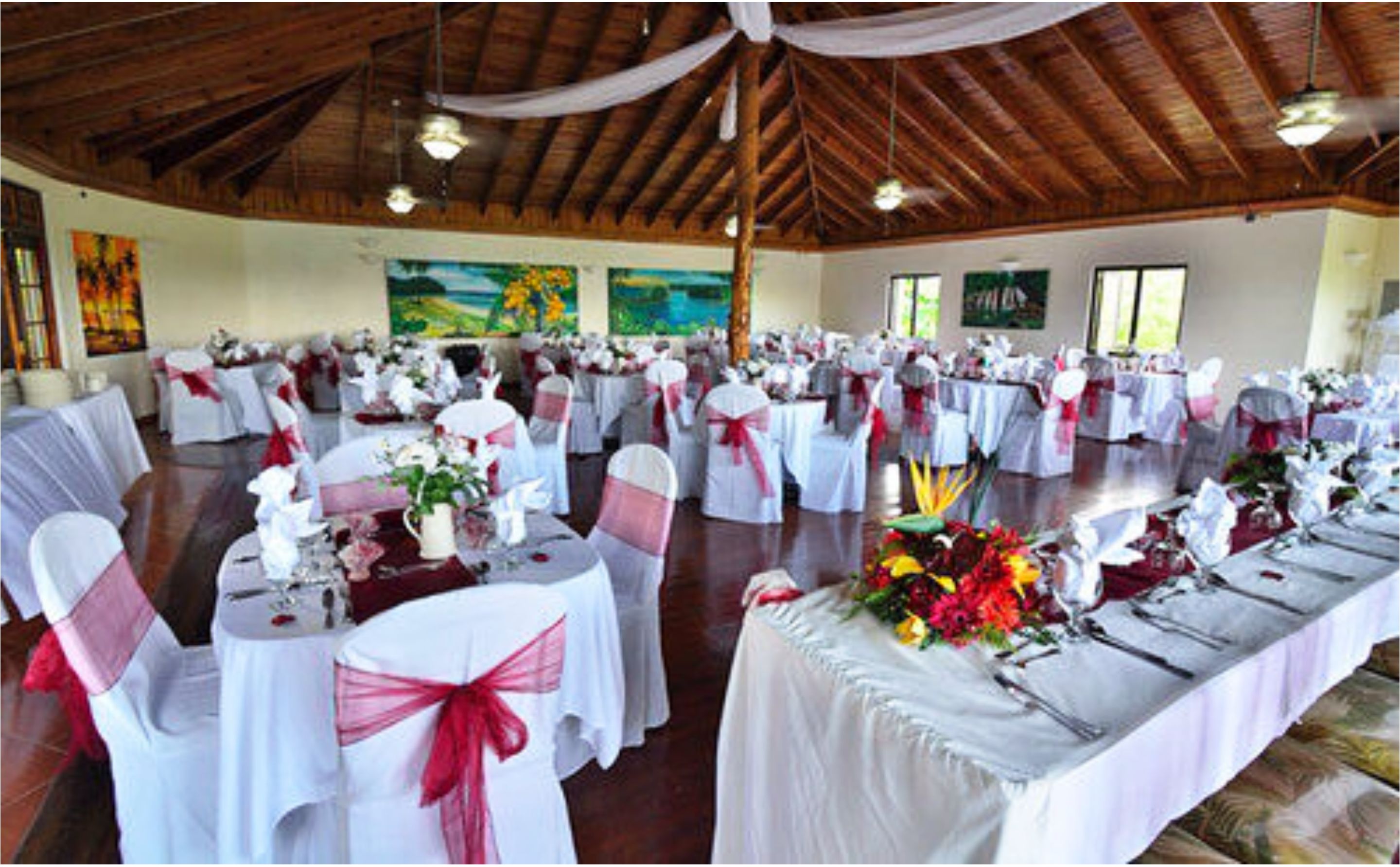 Un salón de banquetes con mesas redondas, manteles blancos y lazos rojos.