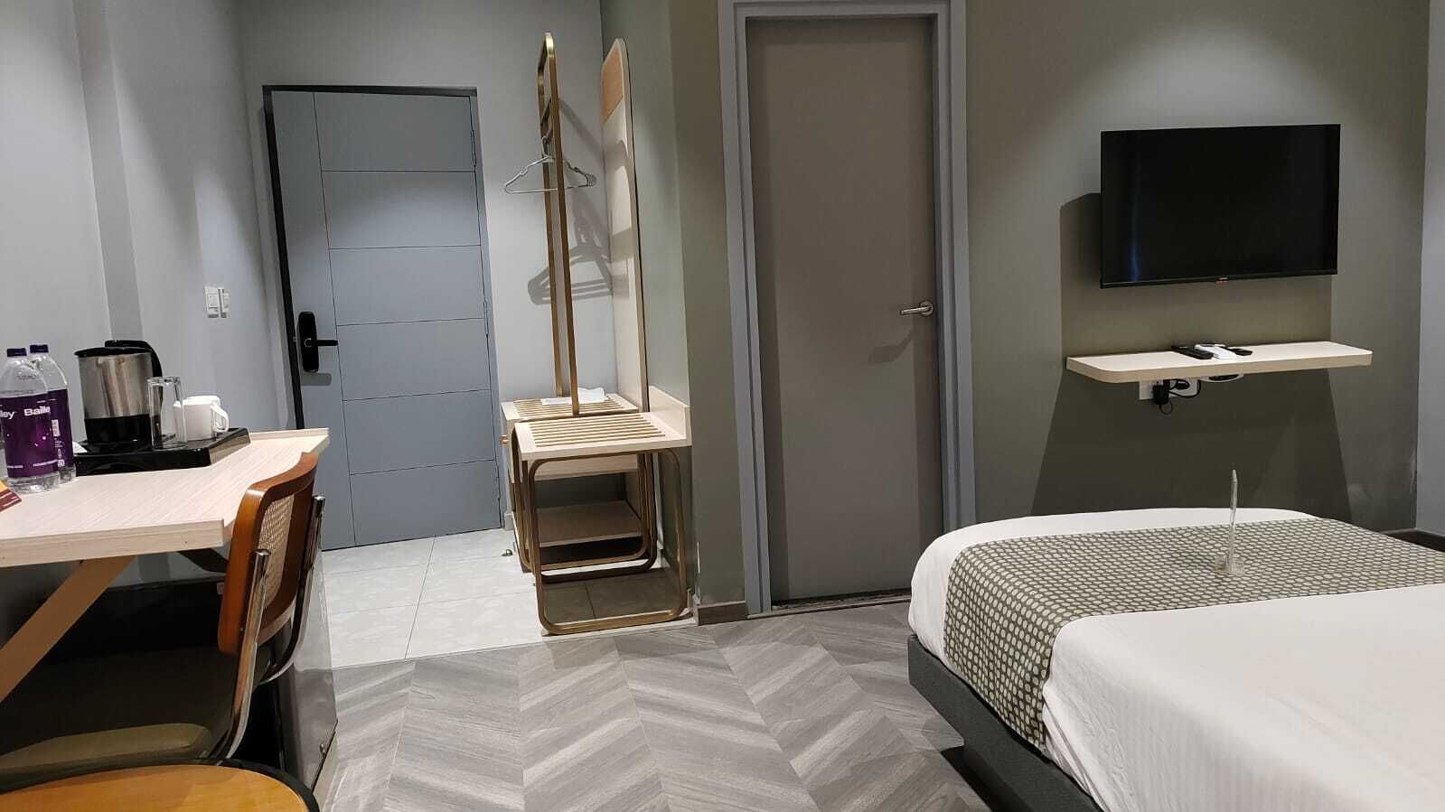 Una habitación de hotel con cama, escritorio con silla, televisor montado en la pared y una puerta.