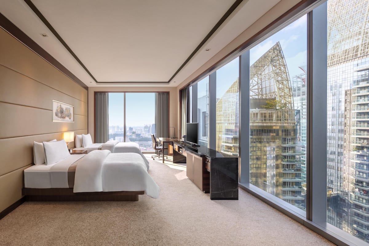 Une chambre d’hôtel moderne dotée d’un grand lit, d’un bureau et d’une télévision. Elle offre une vue sur la ville grâce à de grandes fenêtres.