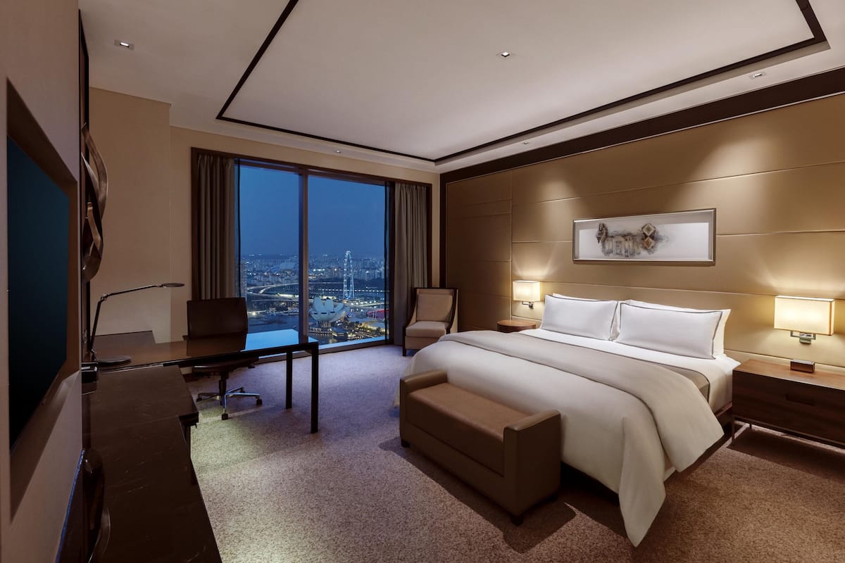 Une chambre d’hôtel moderne, dotée d’un grand lit, d’un bureau et d’une chaise. De grandes fenêtres offrent une vue sur la ville.
