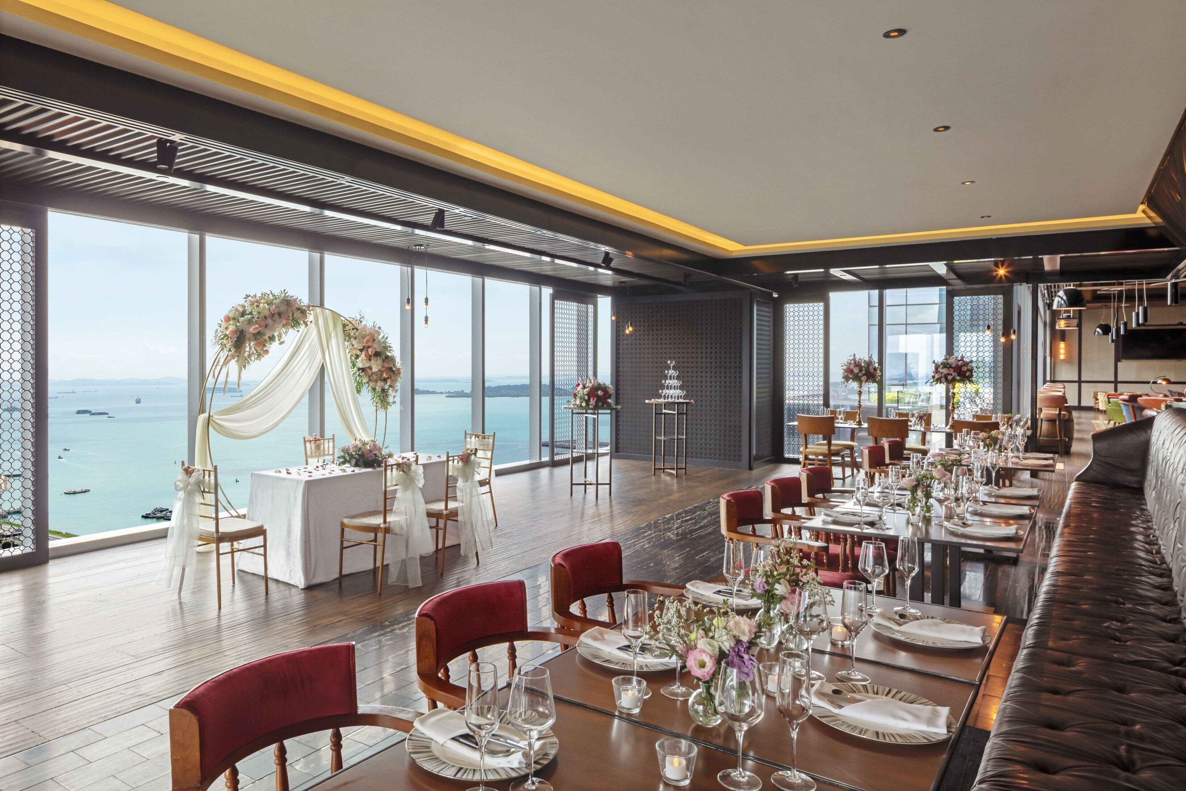 Un espace repas avec des tables dressées pour le repas, de grandes fenêtres offrant une vue sur la mer et un plafond haut agrémenté de luminaires modernes.