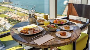 Une table à manger dressée avec des aliments pour le petit-déjeuner, une vue sur la mer et un paysage urbain en arrière-plan.