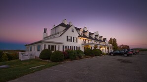 A large, two-story building with a porch and multiple chimneys, surrounded by a parking lot and greenery.