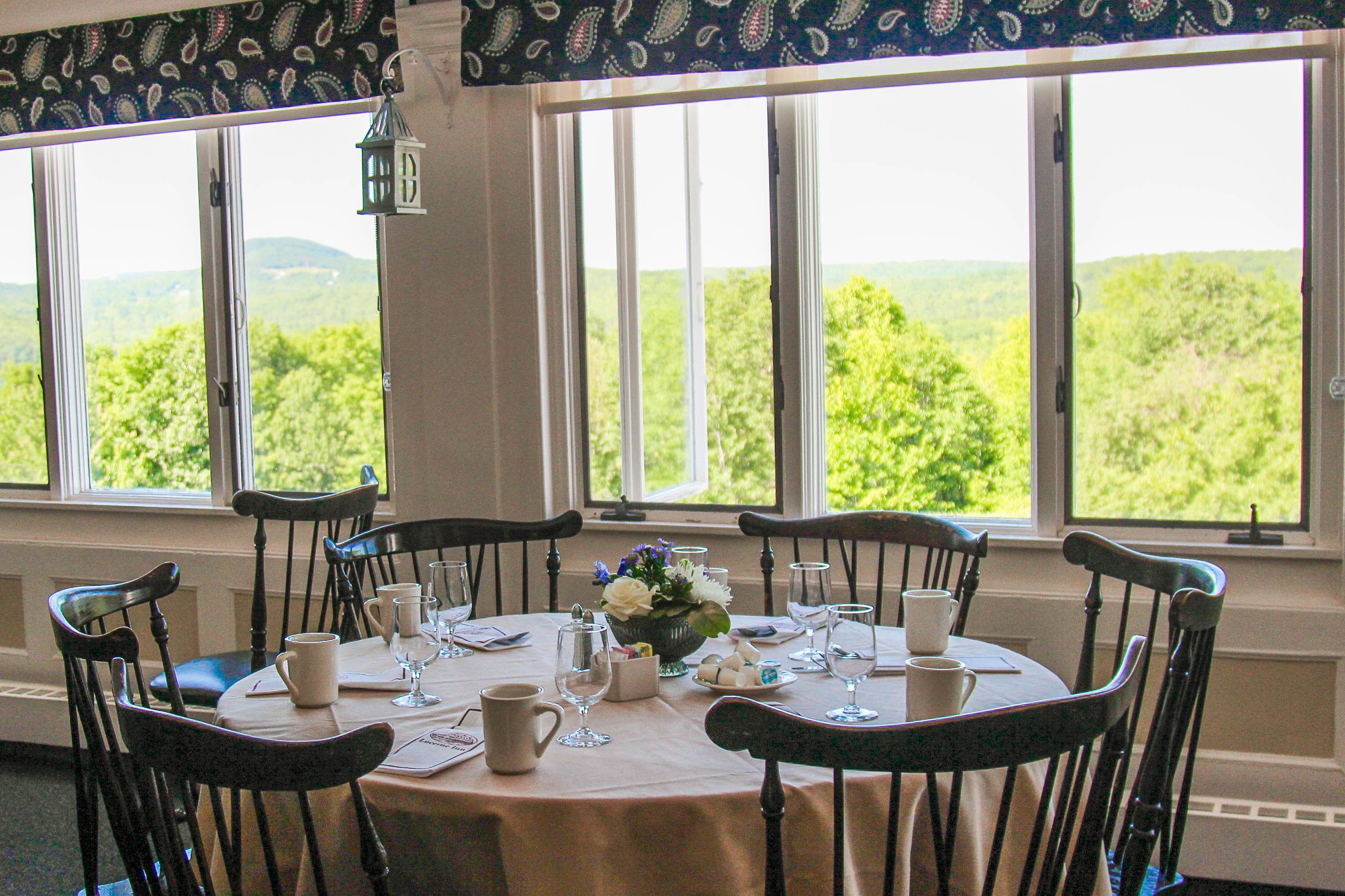 A dining table set for six with chairs, a flower centerpiece, and a view of greenery through large windows.