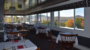 A restaurant with tables and chairs arranged for dining, large windows offering a view of autumn foliage and hills, and a ceiling with a patterned design.