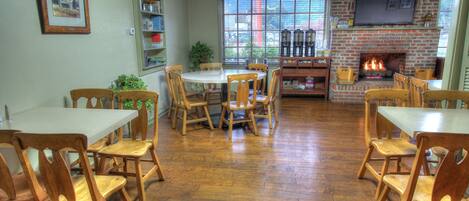 A dining area with wooden tables and chairs, a brick fireplace, and a large window with a view of the outside.