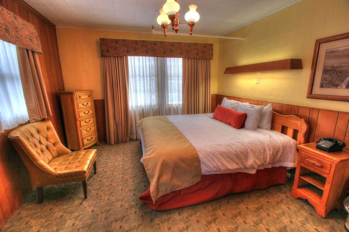 A hotel room with a large bed, a wooden nightstand, a chair, a dresser, and a chandelier.