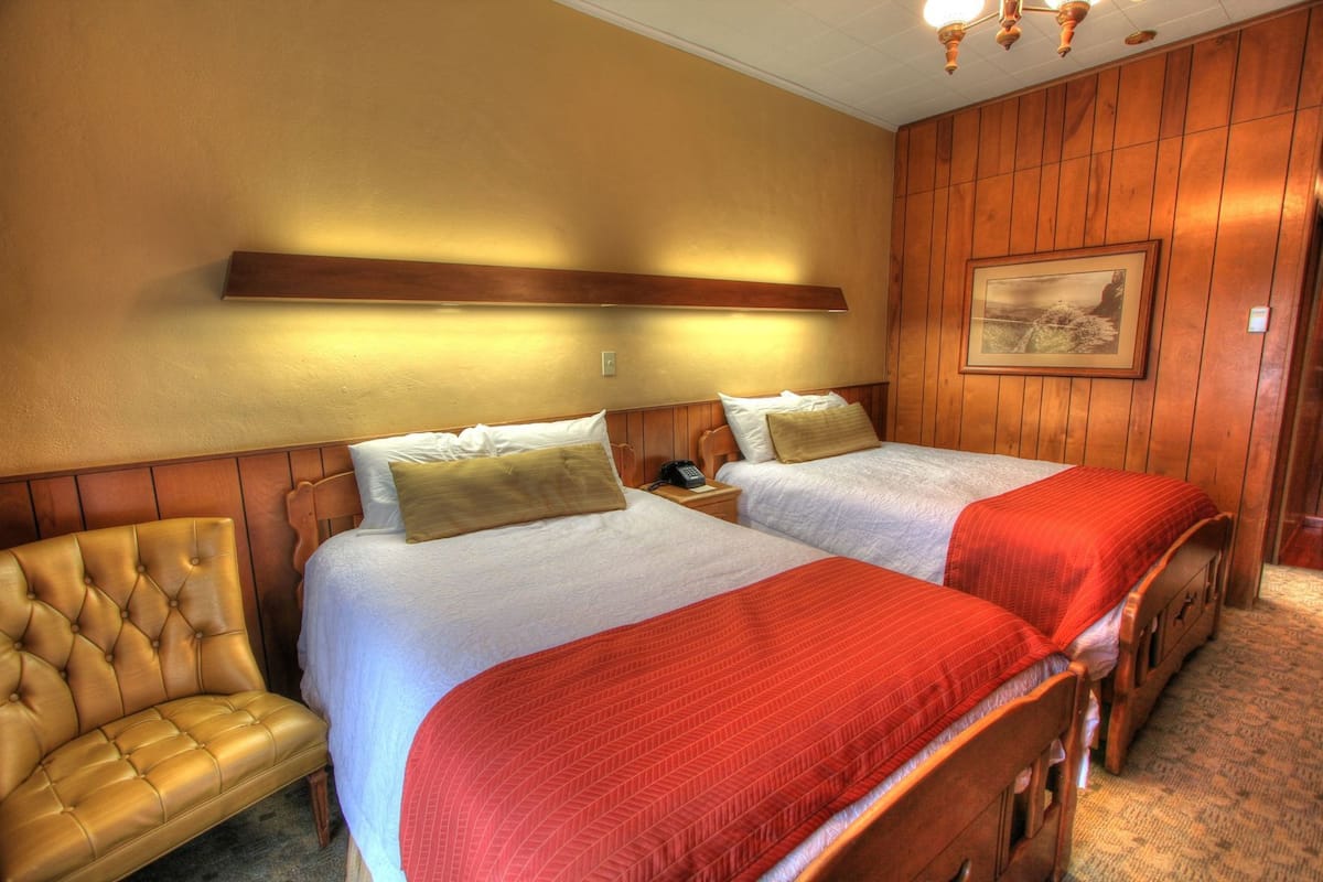 A hotel room with two beds, a leather armchair, wooden paneling, a painting on the wall, and a telephone on the nightstand.