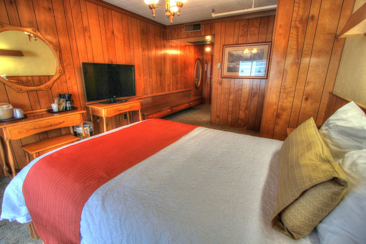 A hotel room with a wooden interior, a large bed with a red and white bedspread, a television, a desk with a coffee maker, and a framed picture on the wall.
