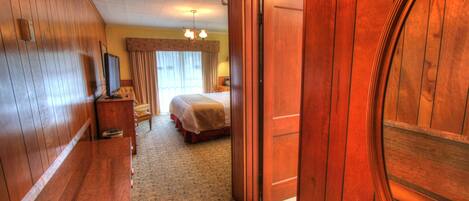 Une chambre d’hôtel avec un plancher en bois, un lit recouvert d’une couvre-lit beige, une table de chevet avec une lampe et une porte en bois.