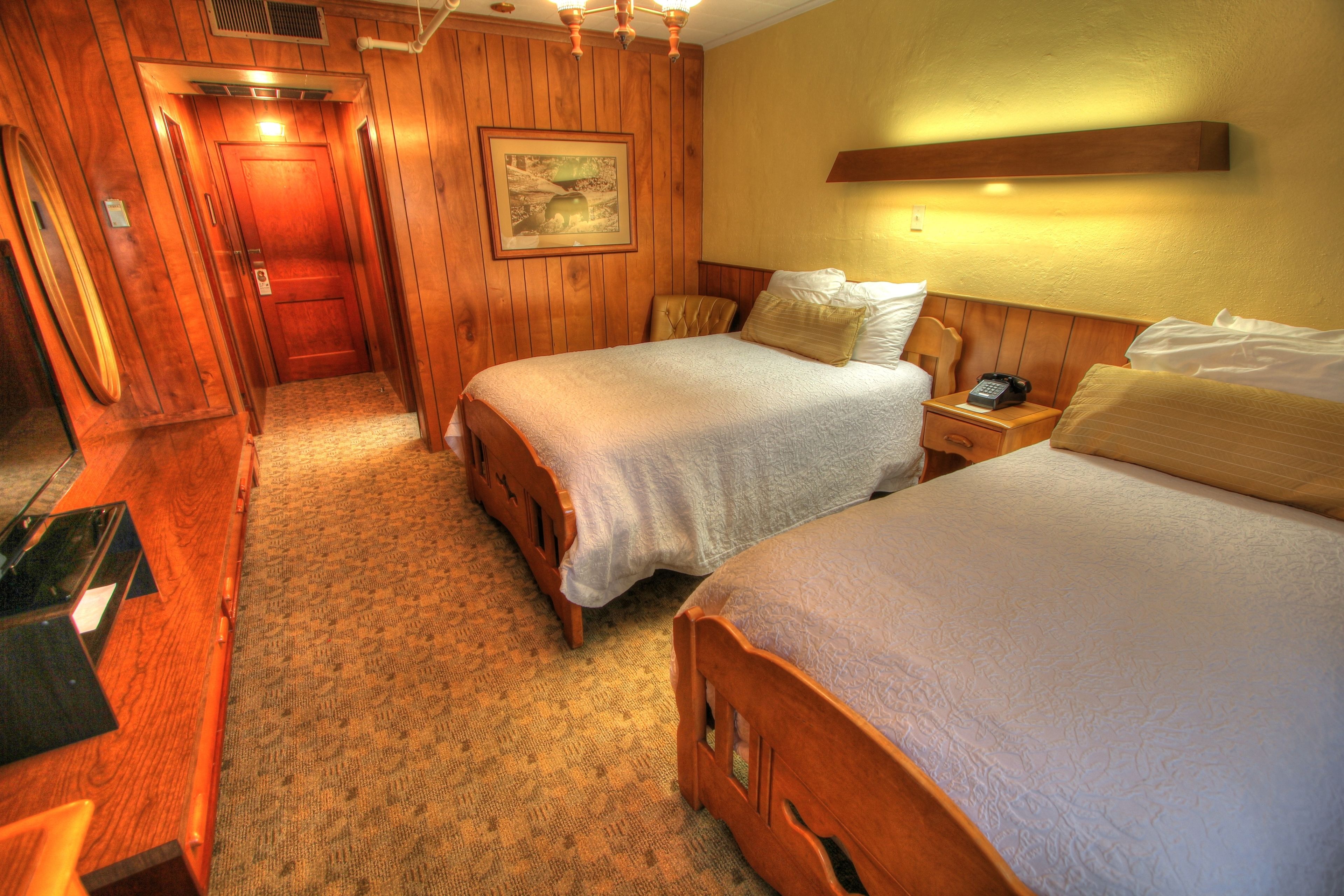 A hotel room with two beds, wooden walls, a carpeted floor, a painting, and a telephone on the nightstand.