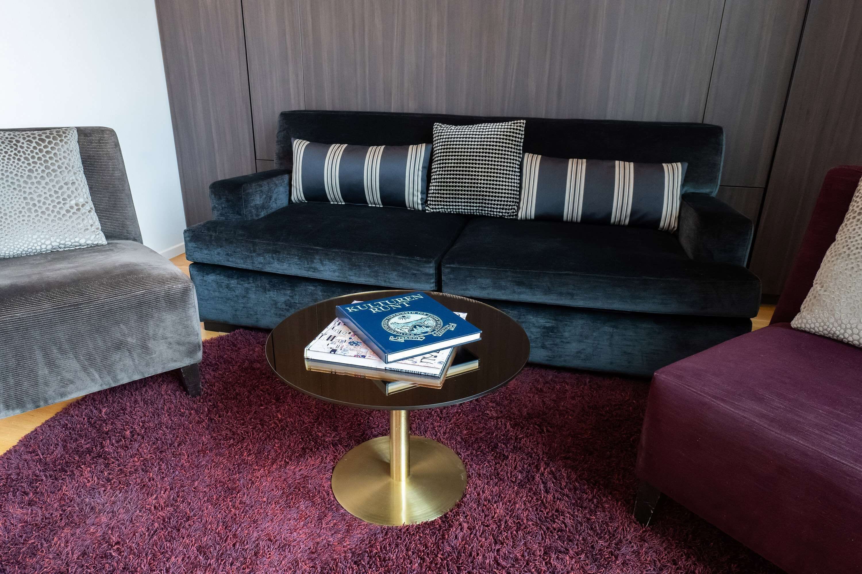 A modern living room with a dark blue sofa, two armchairs, a round coffee table with books, and a red carpet.