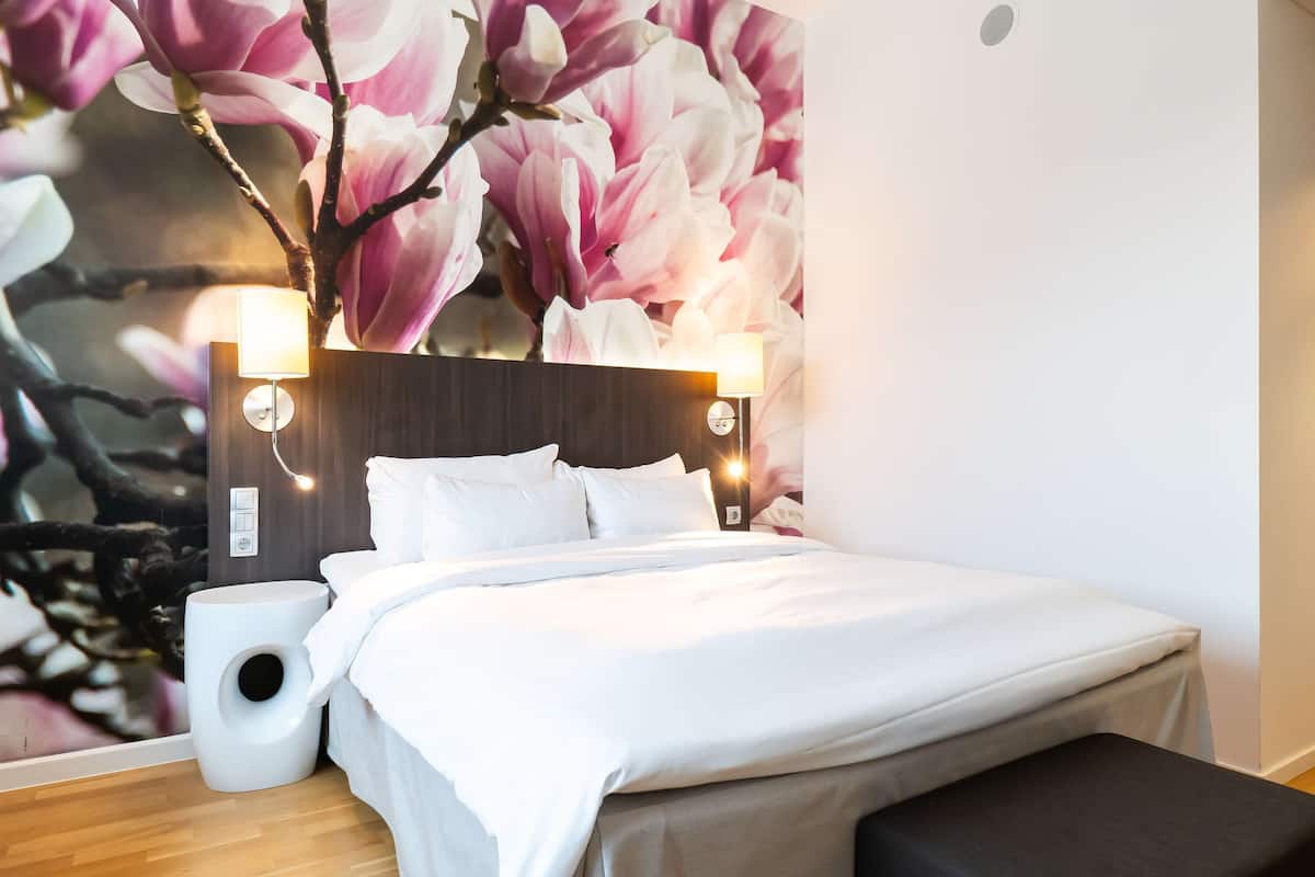 Un dormitorio con un amplio mural floral, cabecera de madera, dos lámparas de noche, colcha blanca y un altavoz blanco pequeño.