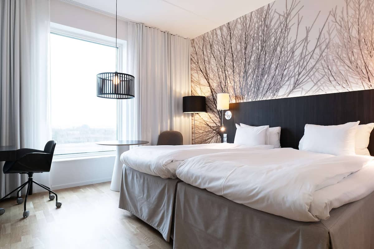 Habitación de hotel con una cama grande, un escritorio y una silla. Hay una ventana con cortinas, una lámpara de pared y una luz de techo.