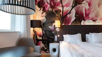A modern hotel room with a large bed, a round table, a chair, a wall-mounted lamp, and a decorative floral mural.