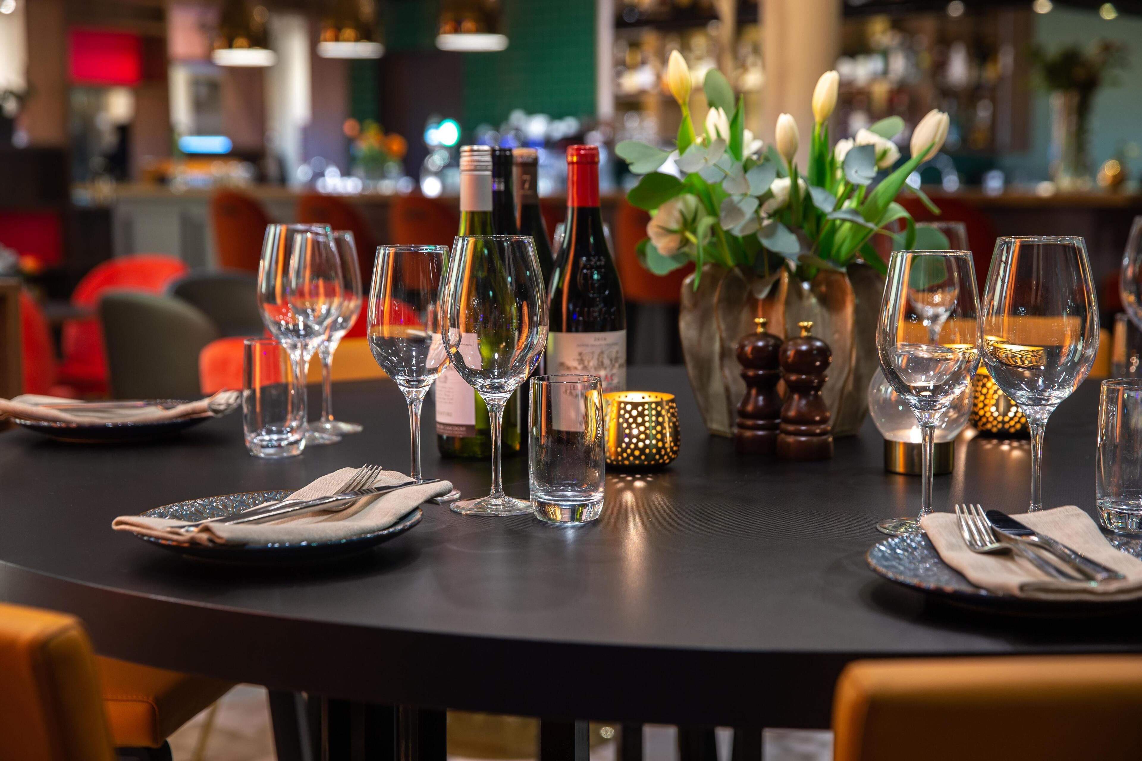 A dining table set with wine glasses, plates, and cutlery, with a vase of flowers and candles in the background.