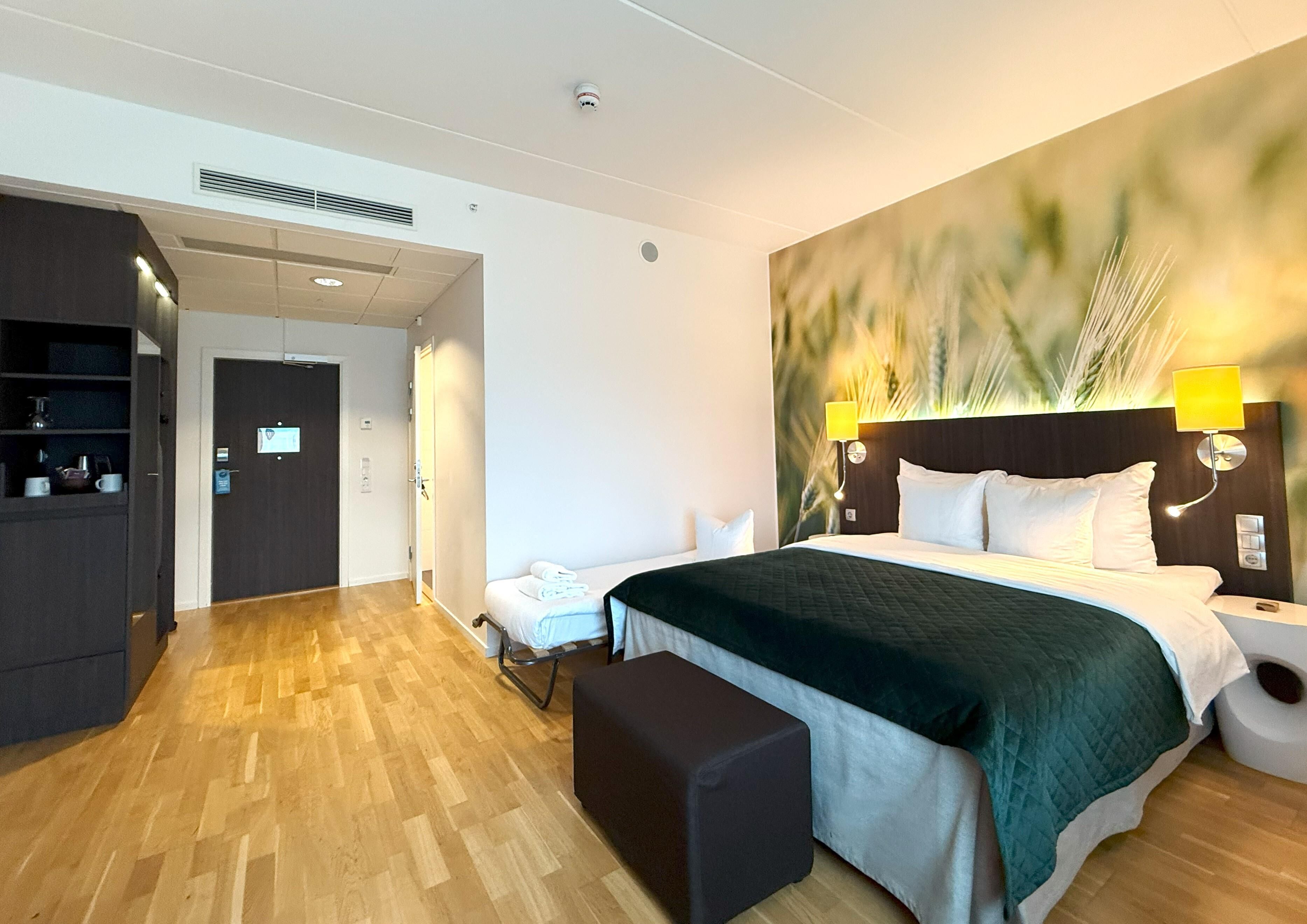 Ett hotellrum med en säng, ett nattduksbord, en lampa och en bänk. Det finns en dörr som leder till en hall med en spegel och en hylla med diverse föremål.