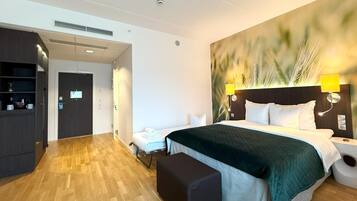 Ett hotellrum med en säng, ett nattduksbord, en lampa och en bänk. Det finns en dörr som leder till en hall med en spegel och en hylla med diverse föremål.