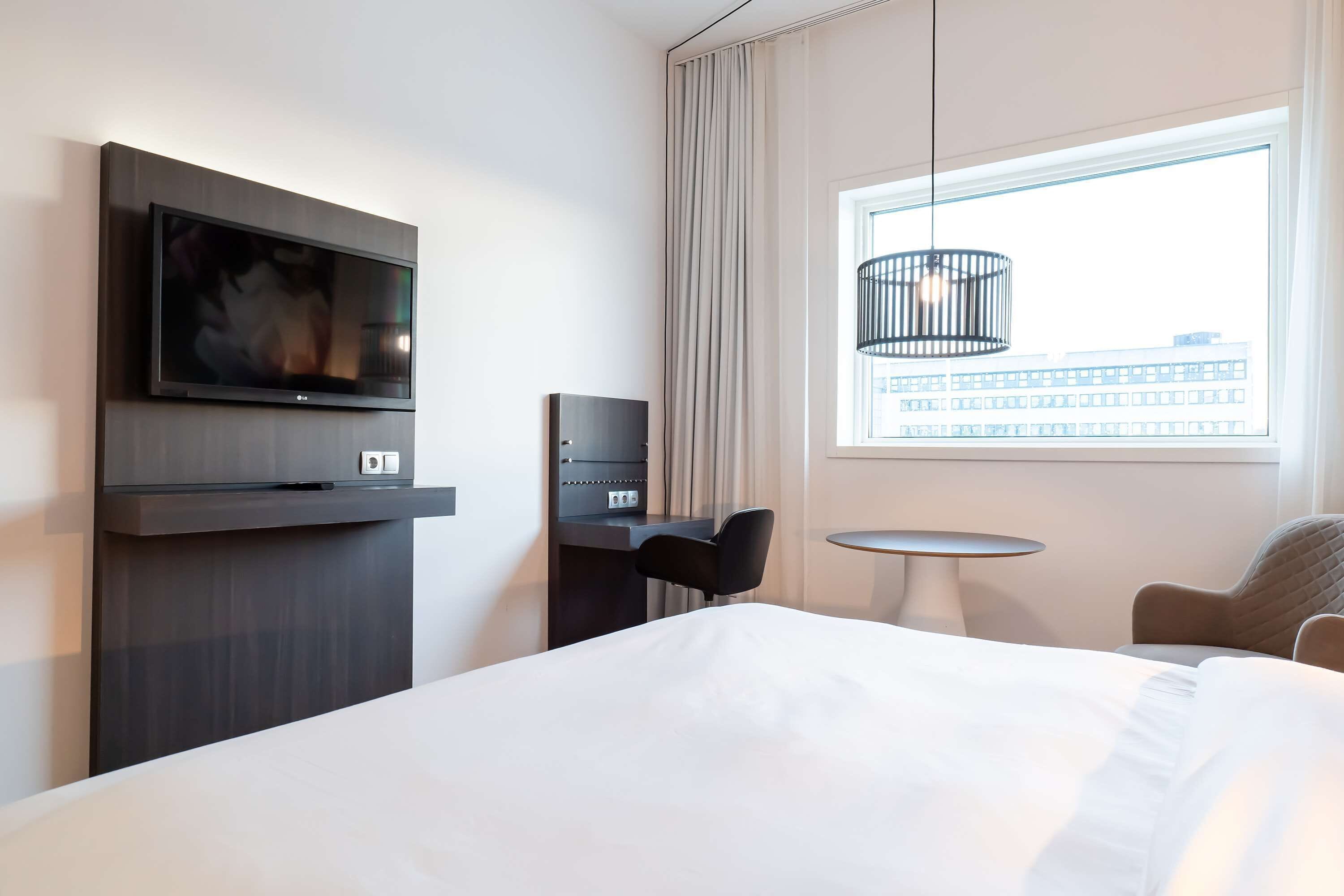 Ein Hotelzimmer mit Bett, Schreibtisch mit Stuhl, Wandfernseher, kleinen Tisch und Fenster mit Blick auf Gebäude.
