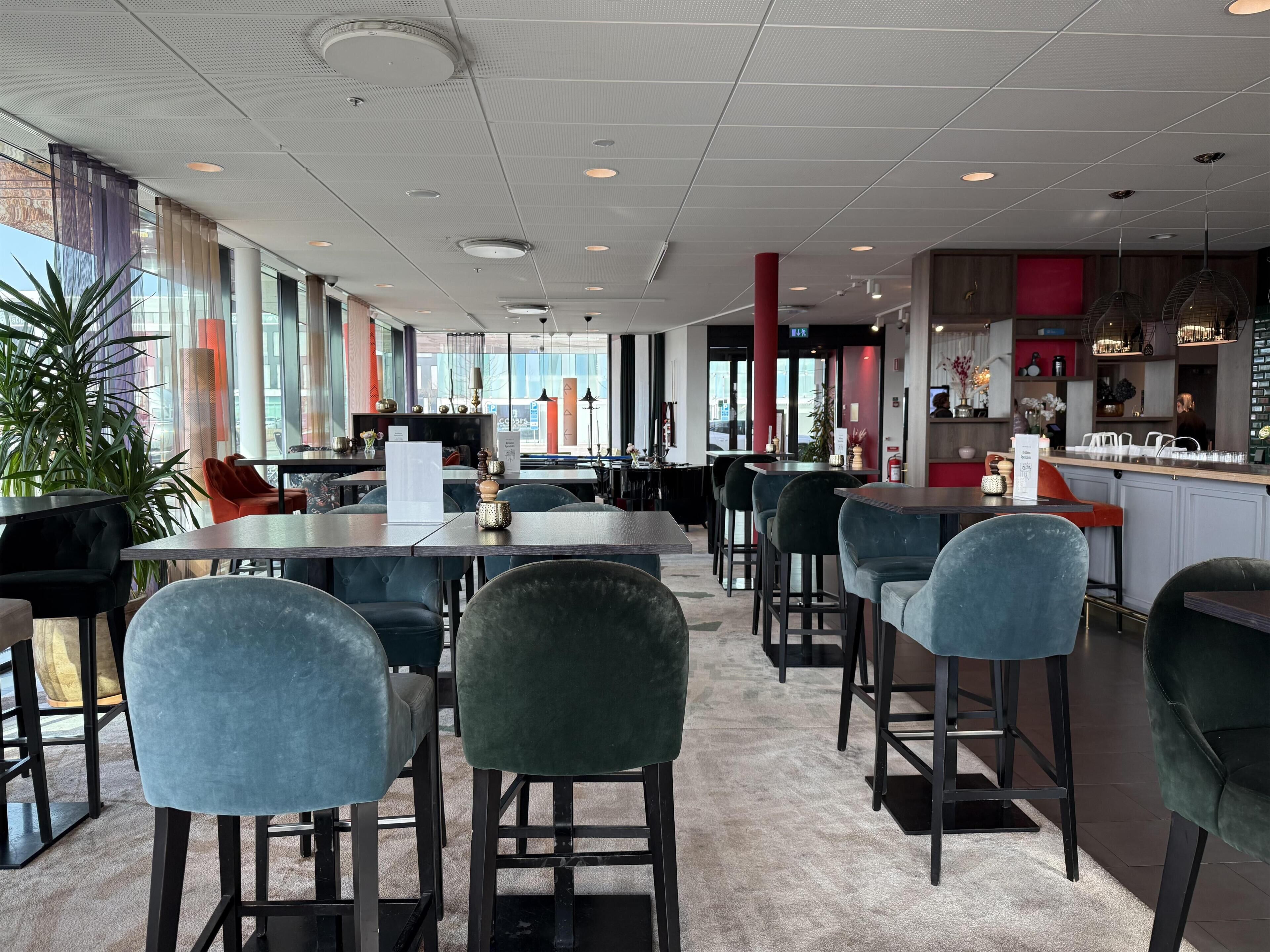 A modern restaurant interior with high ceilings, large windows, and a mix of dining tables and bar seating.