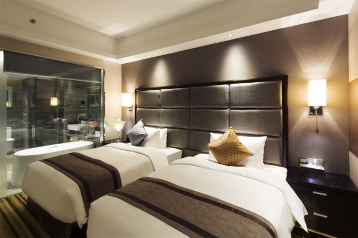 Habitación de hotel con dos camas, un ventanal grande, una bañera y una mesita de noche con lámpara.