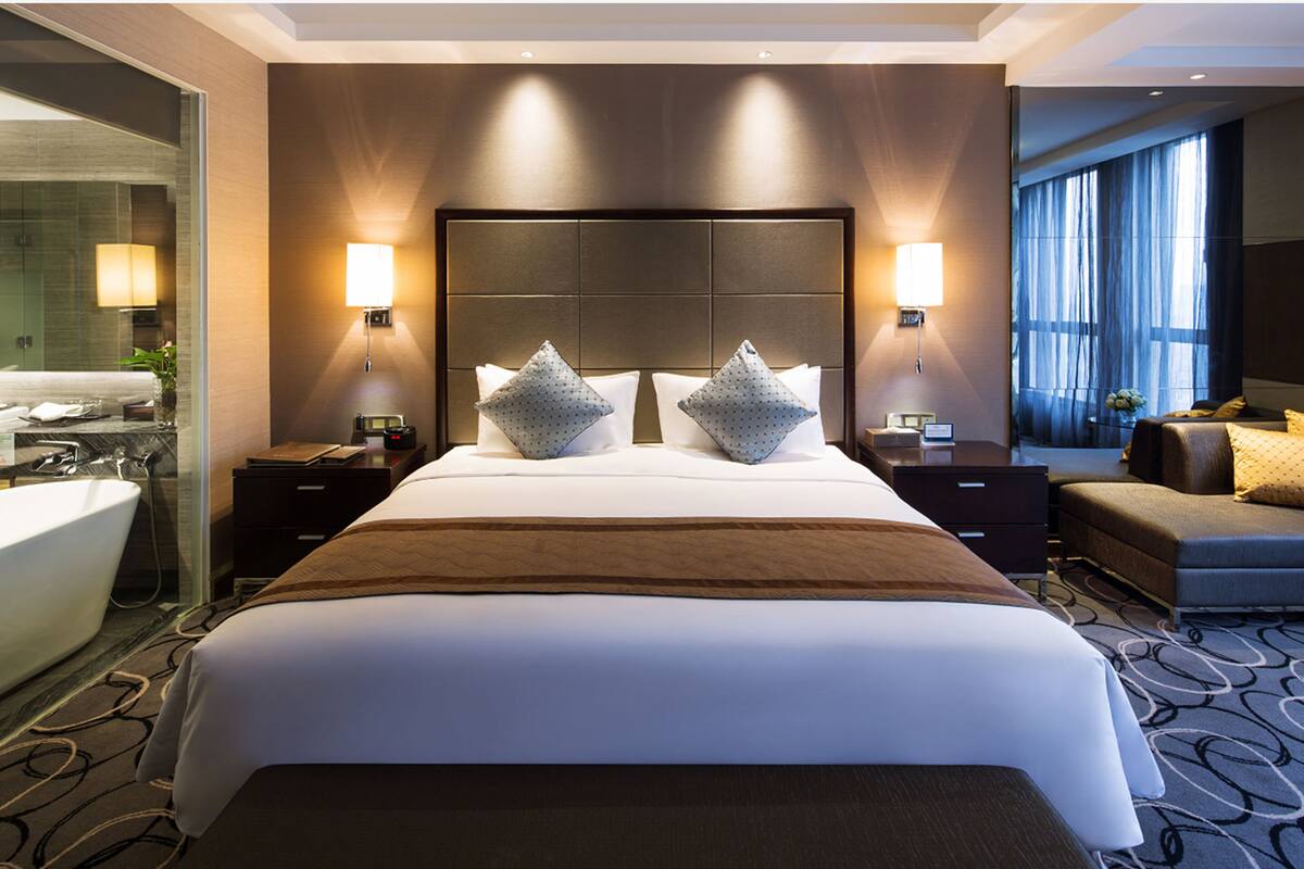 A hotel room with a large bed, two bedside tables, a nightstand, a sofa, a large window, and a bathroom with a bathtub and mirror.