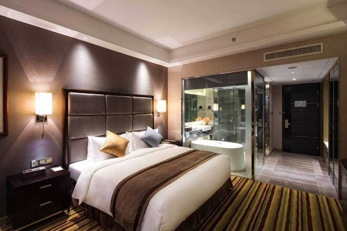 A hotel room with a large bed, bedside tables, a glass partition, and a bathroom visible through the glass.
