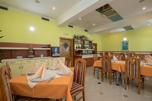 A dining area with multiple tables set for guests, a bar area with shelves of bottles, and a kitchen in the background. - Hotel Adria (Neum)