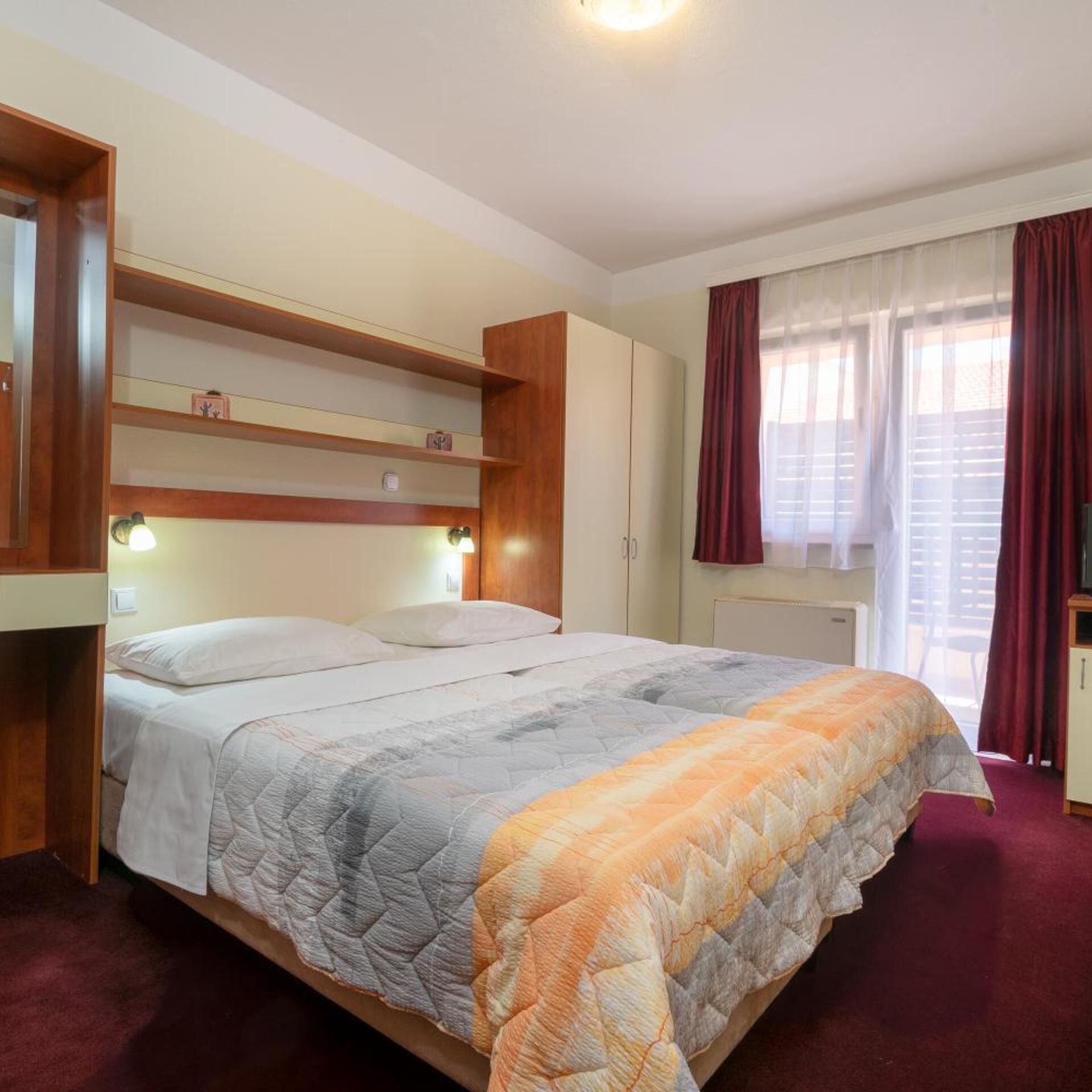 Un dormitorio con cama, cabecera de madera, estanterías empotradas, armario y ventana con cortinas.