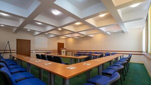 Een vergaderzaal met lange tafels en blauwe stoelen, een spreekgestoelte en een vloerbedekking.