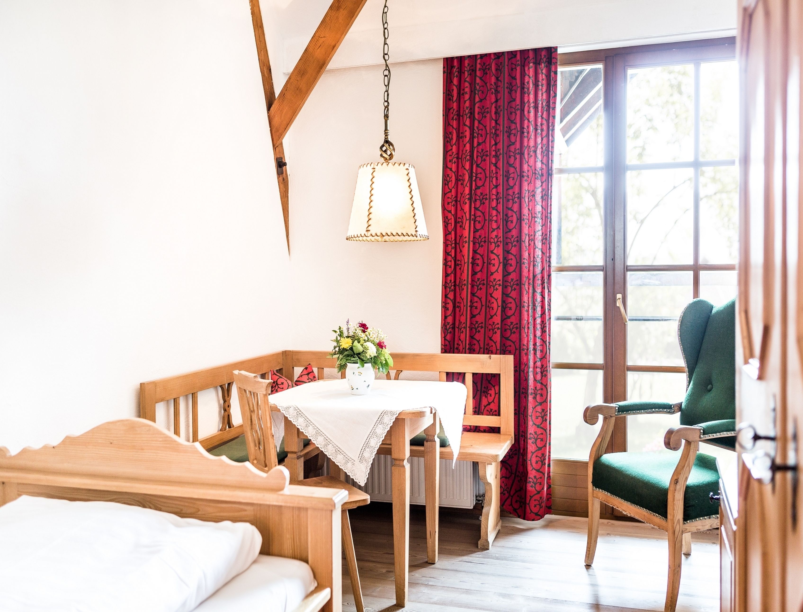 Una habitación acogedora con una cama de madera, una mesa de madera con mantel de encaje, un sillón verde y una ventana con cortinas rojas.